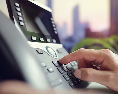 Nahaufnahme eines modernen VoIP-Telefons im Büro: Eine Hand bedient die Telefontastatur, im unscharfen Hintergrund ist eine städtische Skyline zu sehen. Symbolbild für IP-Telefonie, VoIP und professionelle Unternehmenskommunikation.