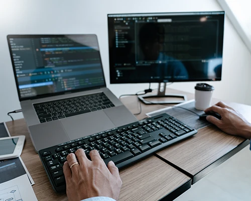 IT-Arbeitsplatz mit Notebook, externem Monitor, Tastatur und Maus: Auf beiden Bildschirmen sind Entwicklungs- und Administrationsoberflächen geöffnet, während eine Person am Computer arbeitet. Symbolbild für IT-Support, Systemadministration und Softwarelösungen.
